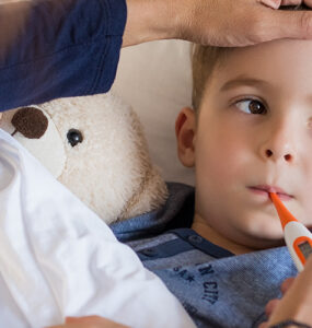 A boy laying in bed, having his temperature taken by his mother to determine if he should go to urgent care of the emergency room.