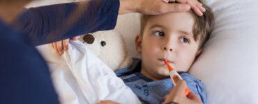 A boy laying in bed, having his temperature taken by his mother to determine if he should go to urgent care of the emergency room.