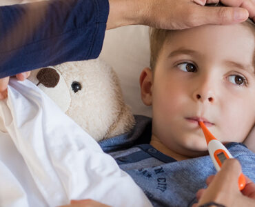 A boy laying in bed, having his temperature taken by his mother to determine if he should go to urgent care of the emergency room.