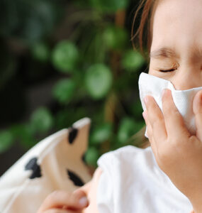 A little girl sitting on the couch, blowing her nose because she has a cold.