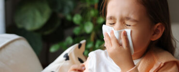 A little girl sitting on the couch, blowing her nose because she has a cold.