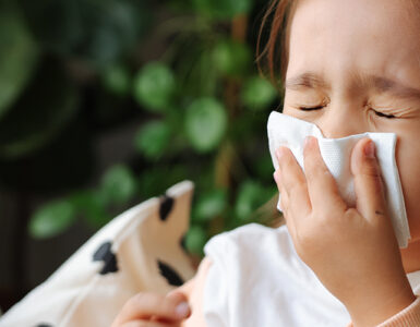 A little girl sitting on the couch, blowing her nose because she has a cold.