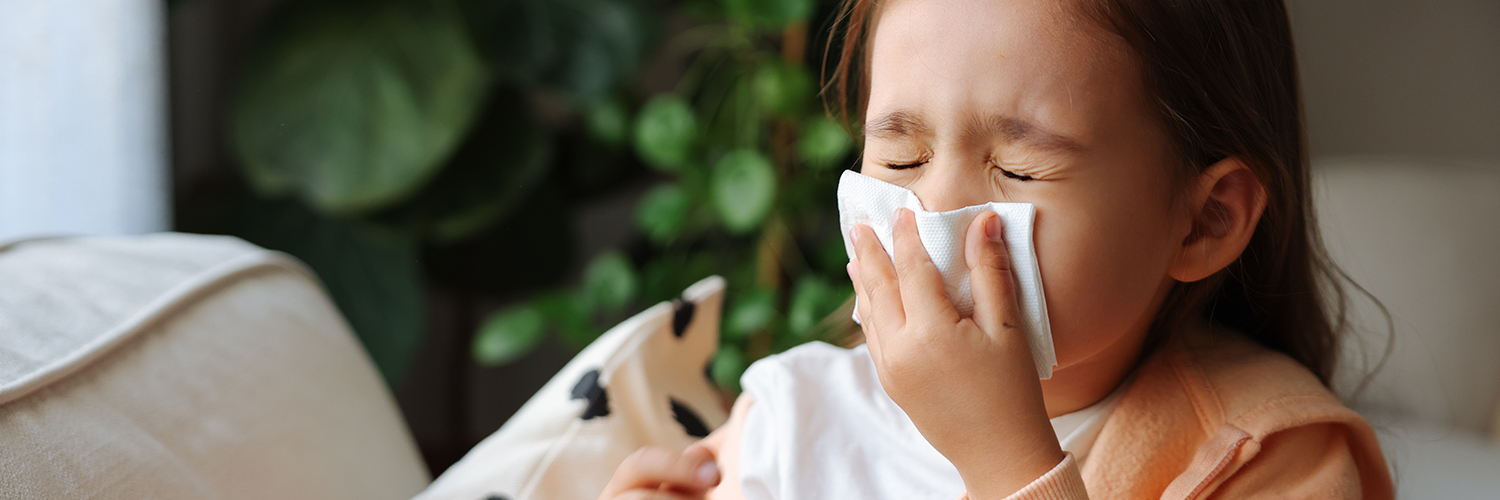 A little girl sitting on the couch, blowing her nose because she has a cold.