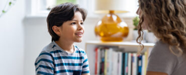 A boy sitting down and looking at his mother, who is noticing his involuntary movements, or tics.