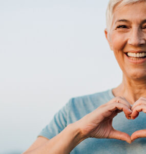 A women outdoors, making a heart shape with her hands and smiling.