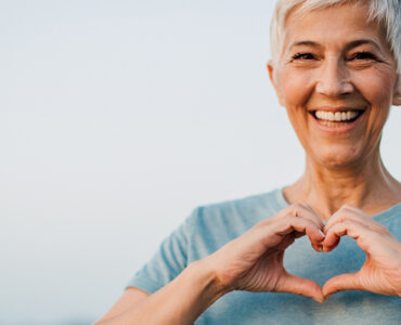 A women outdoors, making a heart shape with her hands and smiling.