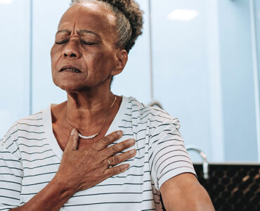 An older woman holding her heart while sitting at her kitchen table.