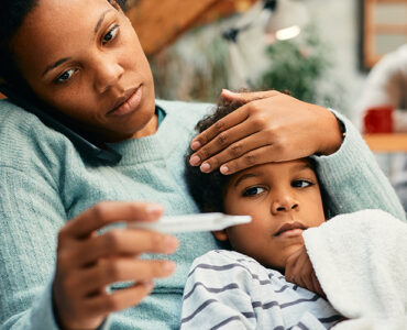 A mother holding her young son, taking his temperature while feeling his forehead.