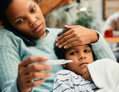 A mother holding her young son, taking his temperature while feeling his forehead.