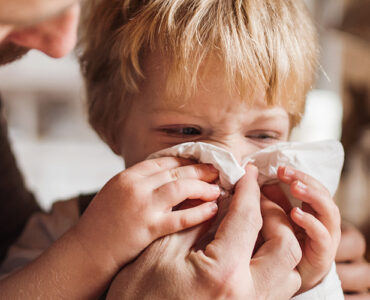 A young boy sneezing into a tissue with help from his father.