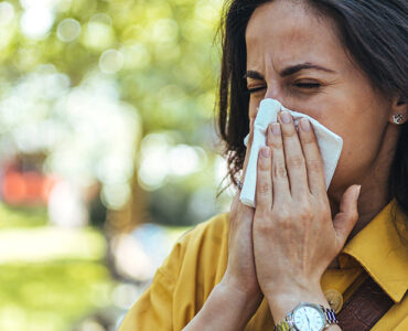 A woman blowing her nose while standing outside with a green tree behind her.