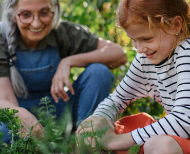 A red-headed young girl gardening with her grandmother, both kneeling while digging into the dirt.