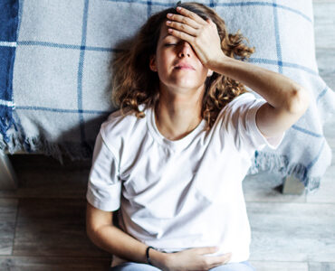 A woman laying down on her bedroom floor, holding her head in pain.
