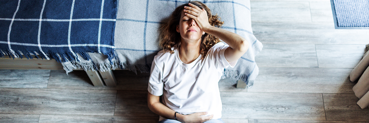 A woman laying down on her bedroom floor, holding her head in pain.