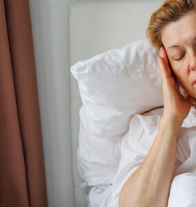 A middle-aged woman with light hair and a white shirt sitting in bed, holding her head in pain.