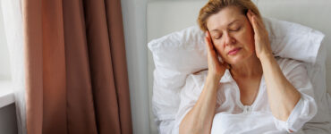 A middle-aged woman with light hair and a white shirt sitting in bed, holding her head in pain.