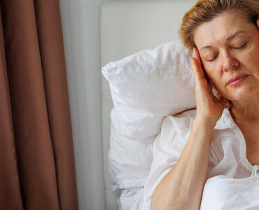 A middle-aged woman with light hair and a white shirt sitting in bed, holding her head in pain.