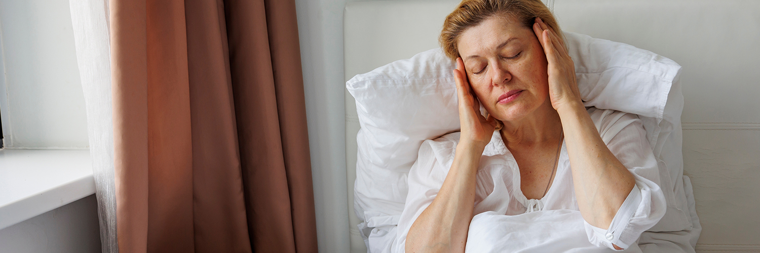 A middle-aged woman with light hair and a white shirt sitting in bed, holding her head in pain.