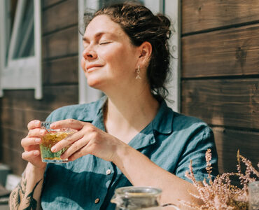 A woman sitting in the sunshine, holding a cup of herbal tea with her eyes closed and a slight smile.