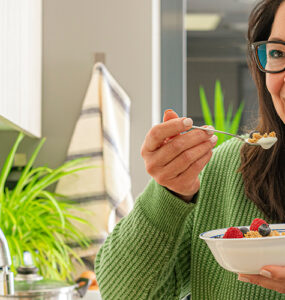 A woman standing in her kitchen, eating a healthy breakfast while smiling.