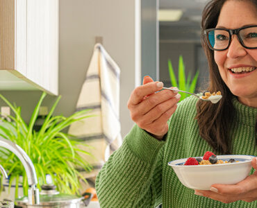 A woman standing in her kitchen, eating a healthy breakfast while smiling.