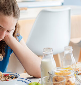 A teenager girl sitting at her kitchen table, playing with her breakfast oatmeal instead of eating it and looking sad.