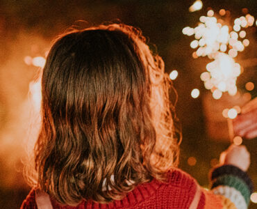 A young girl playing with a sparkler while an adult hand reaches out to help.