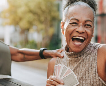 A group of women in their 60s sitting outdoors, playing cards and laughing together.