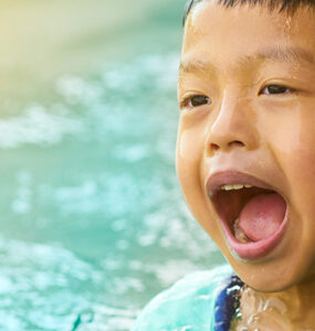 A young boy standing in water, placing his finger in his ear and opening his mouth.