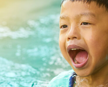 A young boy standing in water, placing his finger in his ear and opening his mouth.