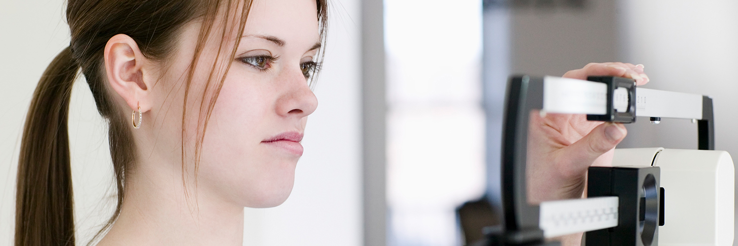 A teenage girl weighing herself on a scale, looking at the results with a frown on her face.