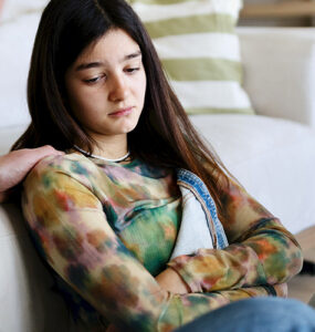 A teenage girl sitting with her father, looking anxious and sad while her father consoles her.