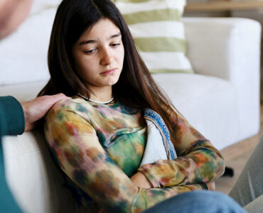 A teenage girl sitting with her father, looking anxious and sad while her father consoles her.