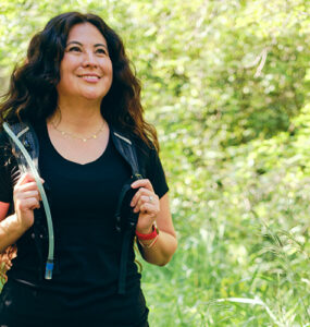 A woman walking outdoors, with greenery behind her, wearing a weighted vest.