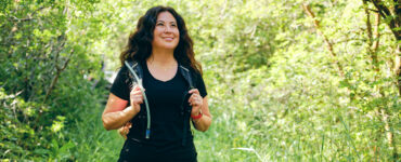 A woman walking outdoors, with greenery behind her, wearing a weighted vest.