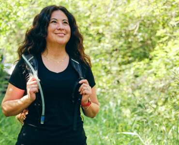 A woman walking outdoors, with greenery behind her, wearing a weighted vest.