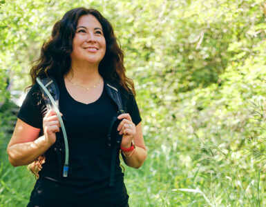 A woman walking outdoors, with greenery behind her, wearing a weighted vest.