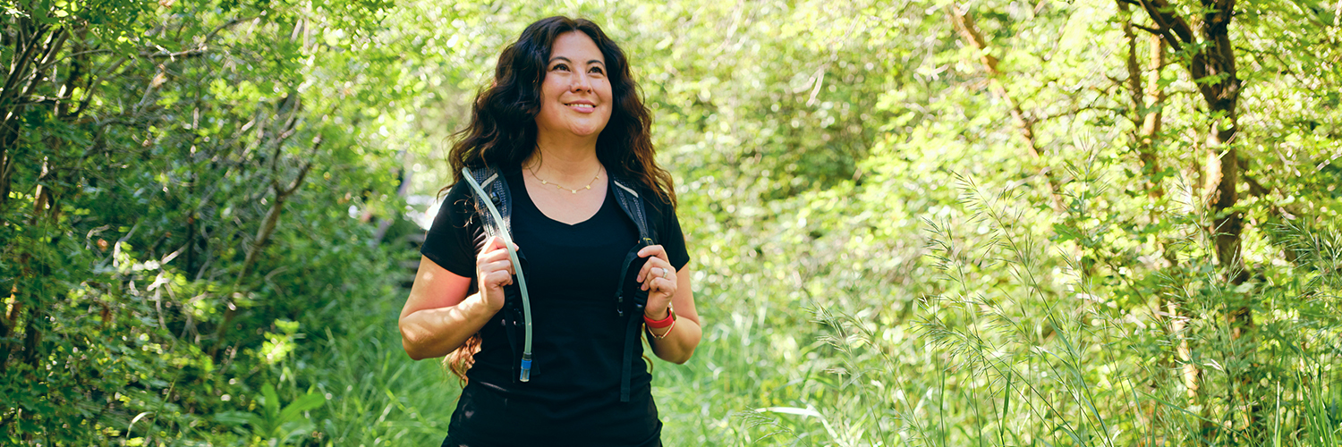 A woman walking outdoors, with greenery behind her, wearing a weighted vest.