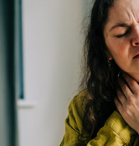A woman sitting on her couch, clutching her throat because it's in pain.