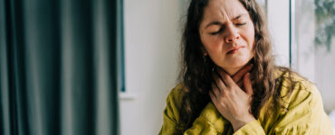 A woman sitting on her couch, clutching her throat because it's in pain.