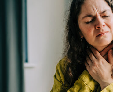 A woman sitting on her couch, clutching her throat because it's in pain.
