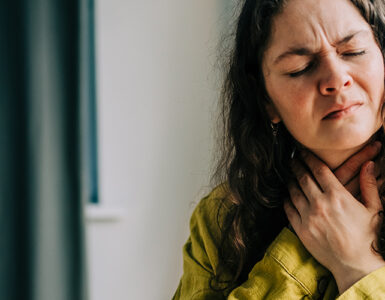A woman sitting on her couch, clutching her throat because it's in pain.