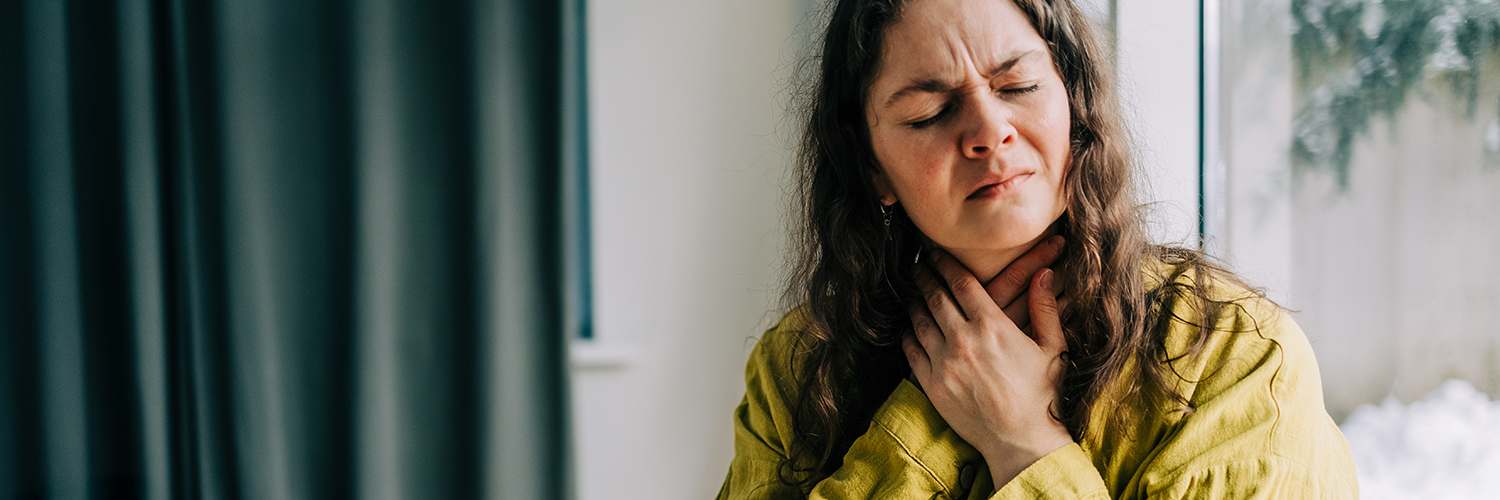 A woman sitting on her couch, clutching her throat because it's in pain.