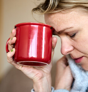 A woman feeling sick with Covid-19 symptoms, holding a tea mug to her forehead with her eyes closed.