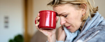 A woman feeling sick with Covid-19 symptoms, holding a tea mug to her forehead with her eyes closed.