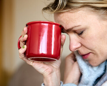A woman feeling sick with Covid-19 symptoms, holding a tea mug to her forehead with her eyes closed.