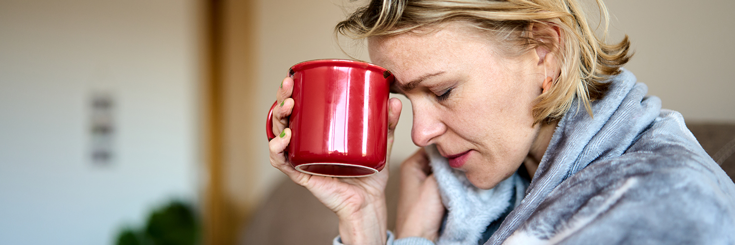 A woman feeling sick with Covid-19 symptoms, holding a tea mug to her forehead with her eyes closed.