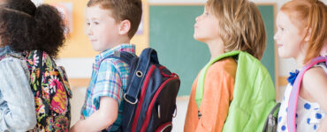 A group of elementary aged students standing in line at school, wearing their backpacks.