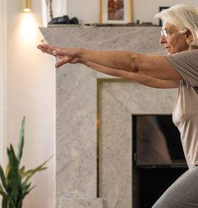 An older woman in her home, exercising on a yoga mat with her hands out in front of her.