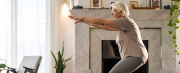 An older woman in her home, exercising on a yoga mat with her hands out in front of her.